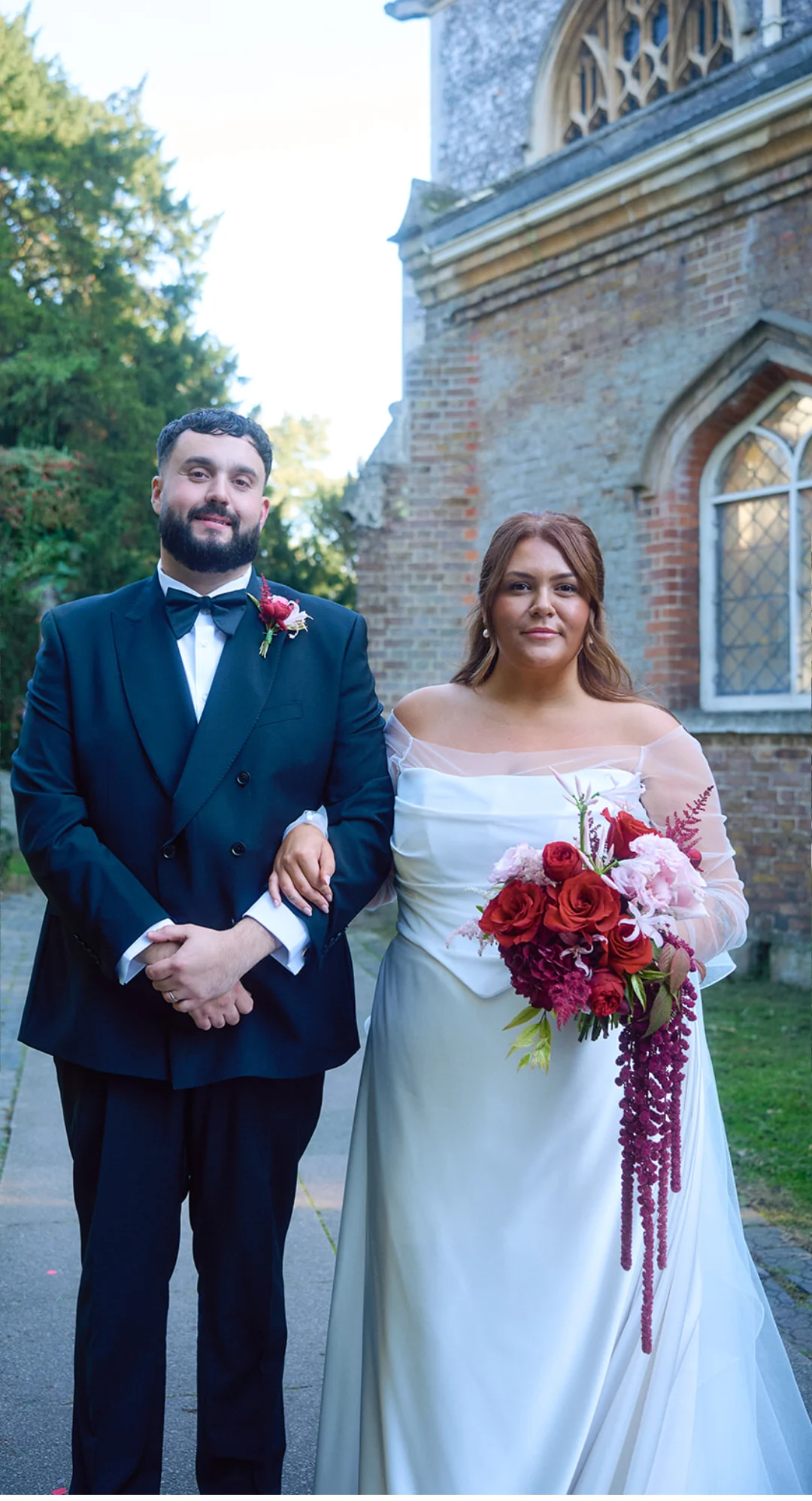 Bride and groom outside their church, the bride in an off-shoulder satin gown with a basque waistline and sheer sleeves, holding a cascading red bouquet