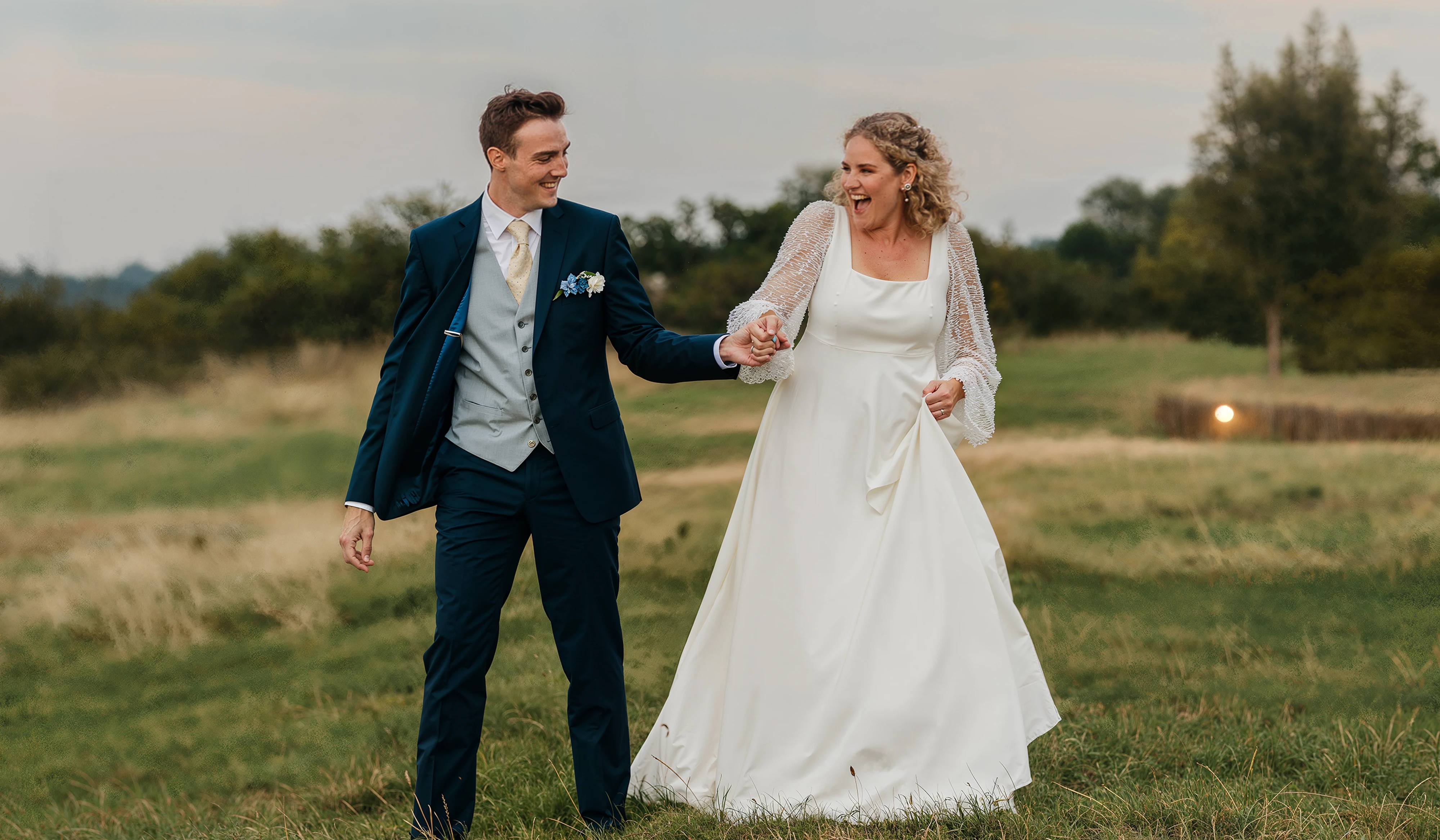 Bride in a satin wedding dress with sheer beaded sleeves walking hand in hand with her groom across a meadow at dusk