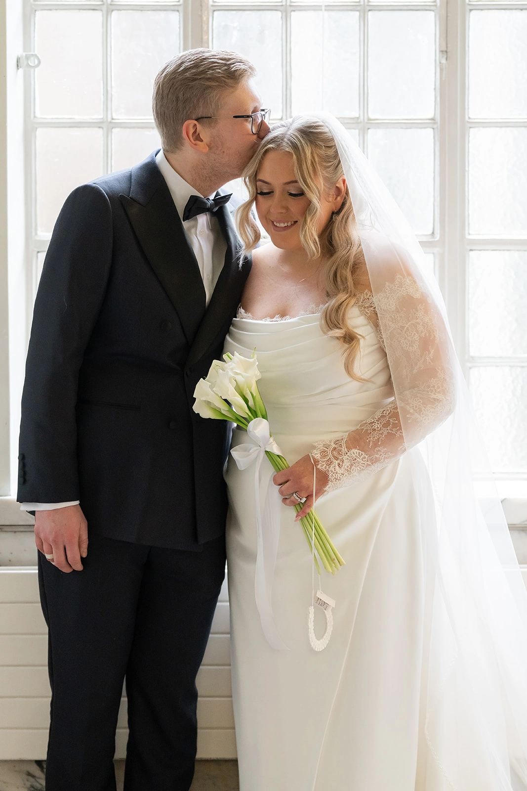 Custom wedding dress with sheer lace sleeves, bride and groom by the window with her cathedral veil