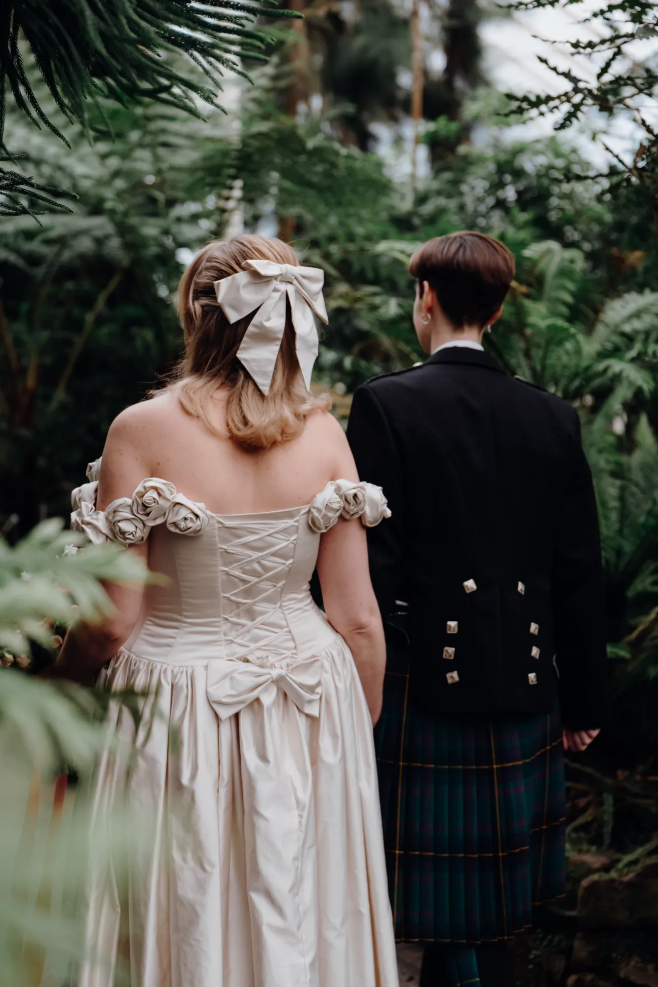 Back view of Flo’s tea-length ballgown with corset lacing, rosette sleeves and a satin bow, walking beside her partner in a kilt