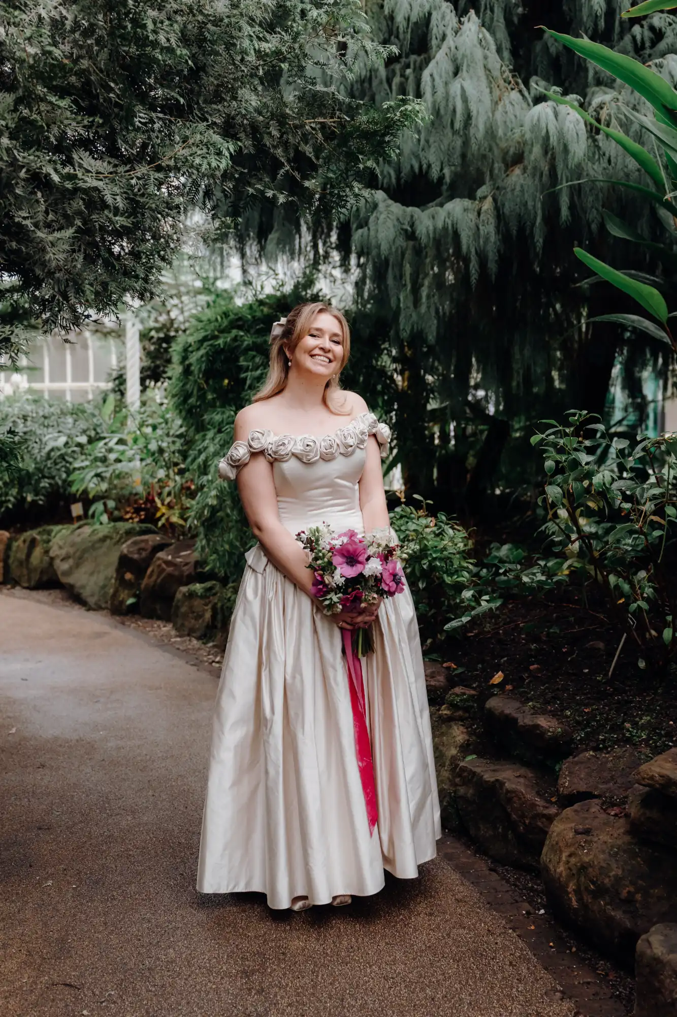 Custom champagne silk duchess satin tea-length ballgown with a rosette off-the-shoulder neckline, front view with a pink anemone bouquet in the glasshouse