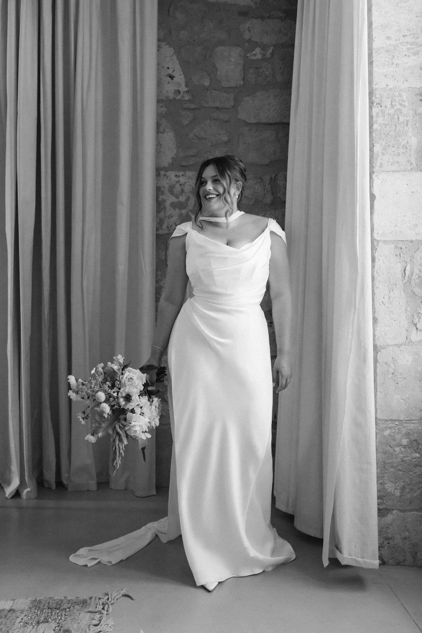 Custom bias-cut satin wedding dress with hidden corsetry, full-length view in the doorway with her bouquet