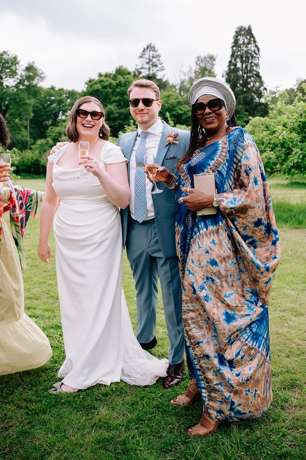 Georgina laughing with her groom and a guest on the lawn, in a draped satin wedding gown with cap sleeves