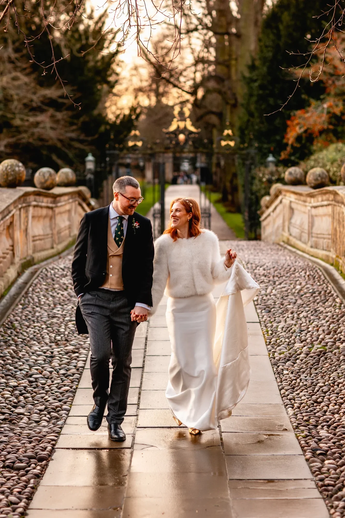 Hermione and her groom walking a cobbled garden path in winter light, her satin wedding skirt held in one hand over a soft white jumper