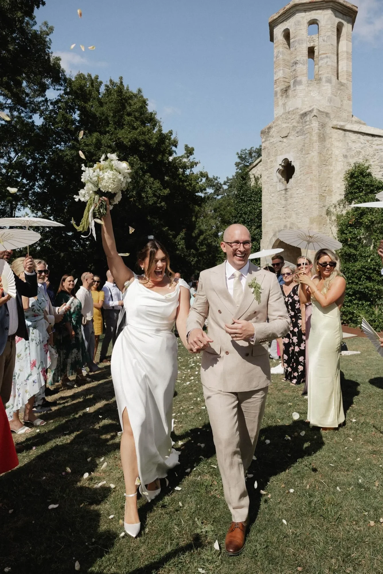 Krystie and her groom walking through petals outside a stone chapel, her draped satin gown caught mid-stride with a side split
