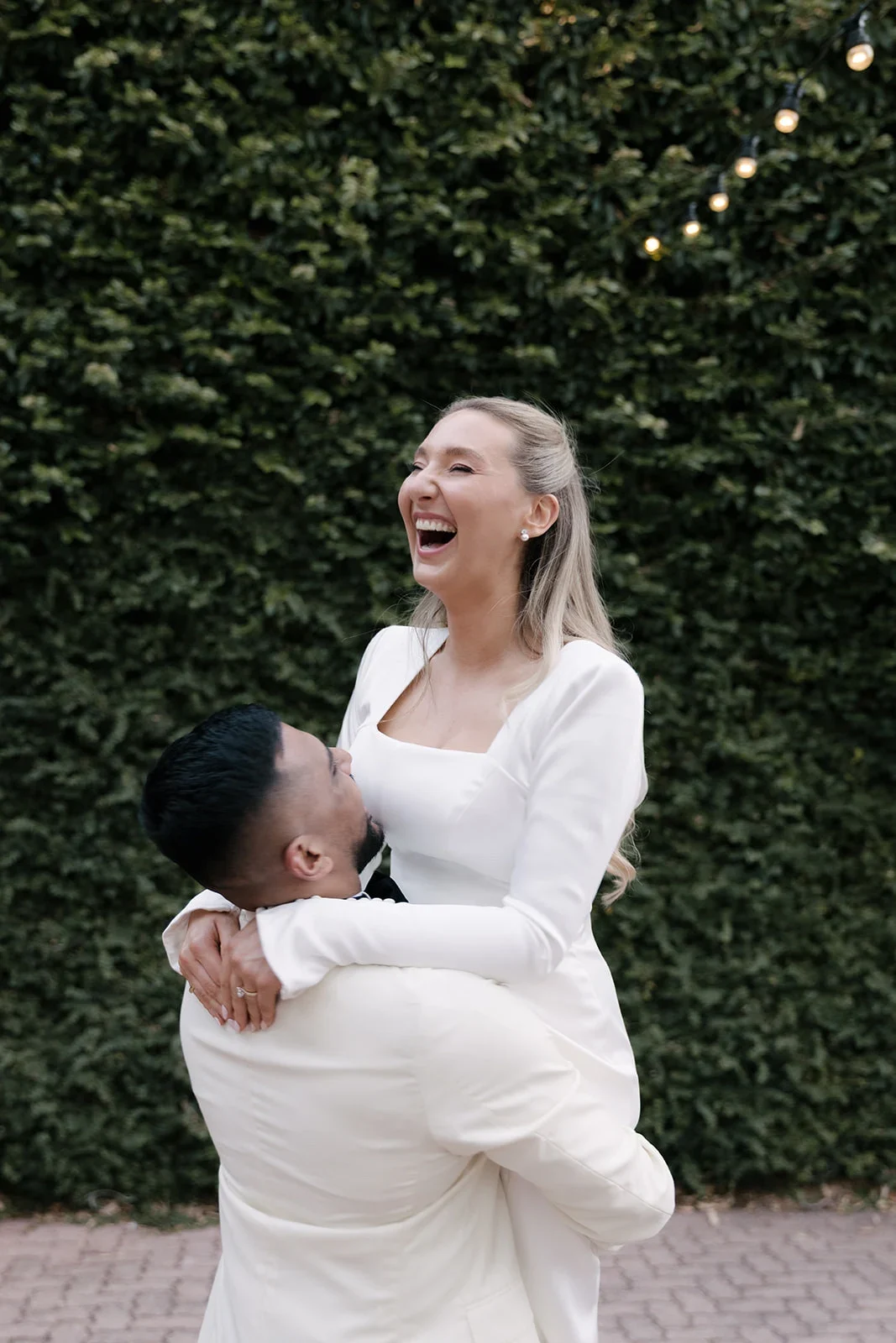 Bride laughing as her groom lifts her, wearing a square-neck crepe gown with long flared sleeves